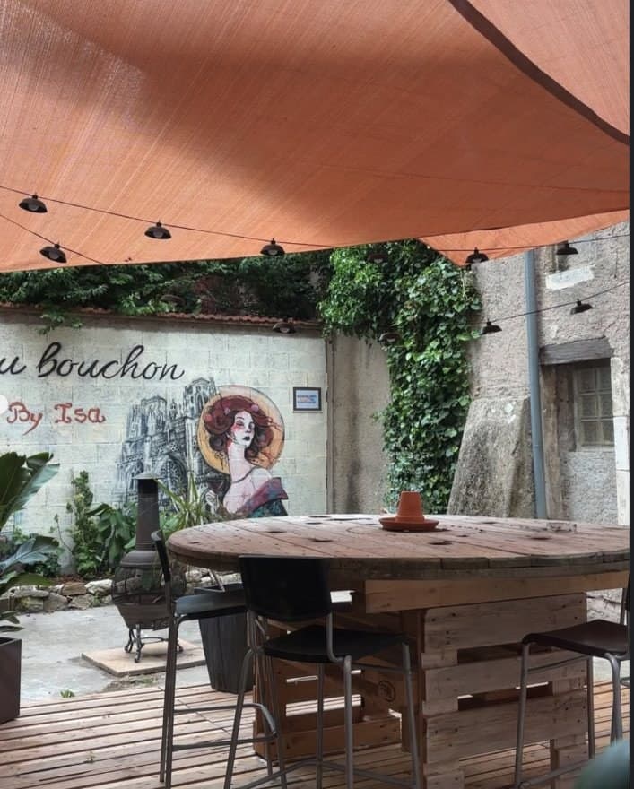 La terrasse d'Au Bouchon by Isa : grande table en touret sous un voile orange, devant la fresque à la femme au chapeau rouge